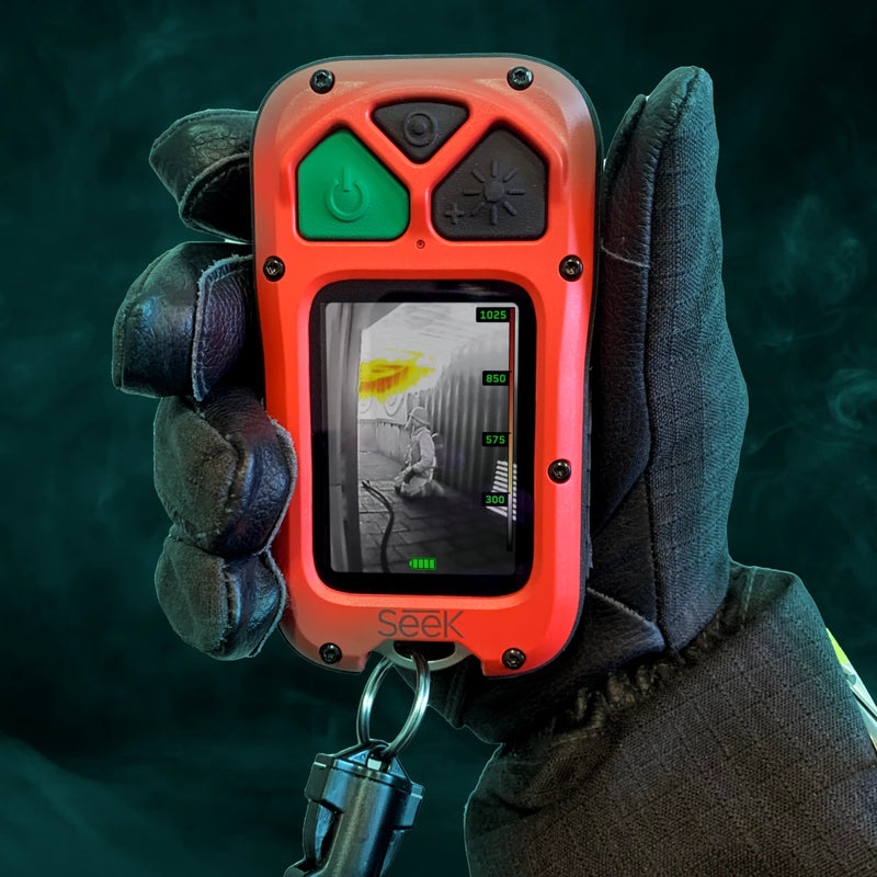 A firefighter holding a red and orange Seek handheld gas detector with a screen showing a gas reading, wearing a black protective glove against a dark, smoky background.