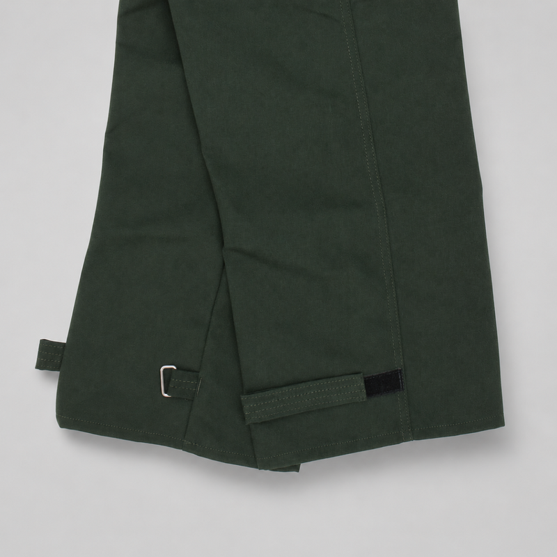 Close-up of spruce green trousers showing the hem with adjustable hook-and-loop strap closures on a light gray background.