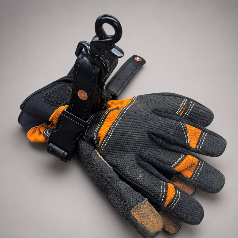 A pair of black and orange work gloves with a black leather wrist strap and metal hook attachment, displayed on a neutral gray background.