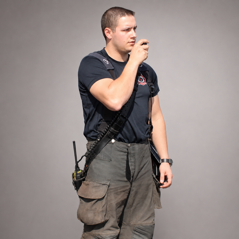 A man wearing a black tactical utility vest with a shoulder strap and a radio pouch, paired with dark cargo pants, standing against a solid gray studio background.