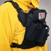 A close-up of a firefighter wearing a yellow turnout jacket and a black tactical chest rig pouch. The pouch has multiple zippered compartments, velcro panels, and a patch.