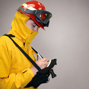 A firefighter in a bright yellow protective suit and helmet with goggles, writing on a clipboard against a plain gray background.