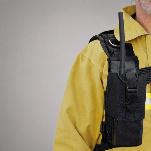 A close-up of a firefighter wearing a yellow turnout jacket and a black tactical vest pouch holding a black handheld radio with an antenna, against a plain gray studio background.