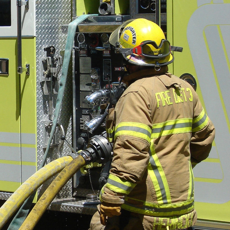 Firefighter connecting a hose to the pump panel on a yellow fire engine during operations.