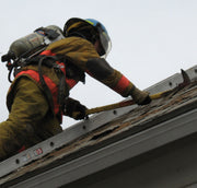 Firefighter wearing turnout gear and SCBA using a pick-head axe on a roof beside a ladder.
