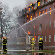 Three firefighters in full turnout gear using hoses to spray water onto a large brick building engulfed in flames during a structure fire.