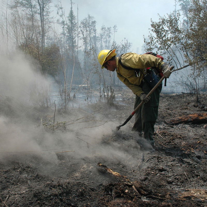Wildland firefighter working with a hand tool on smoky, burned ground during mop-up.